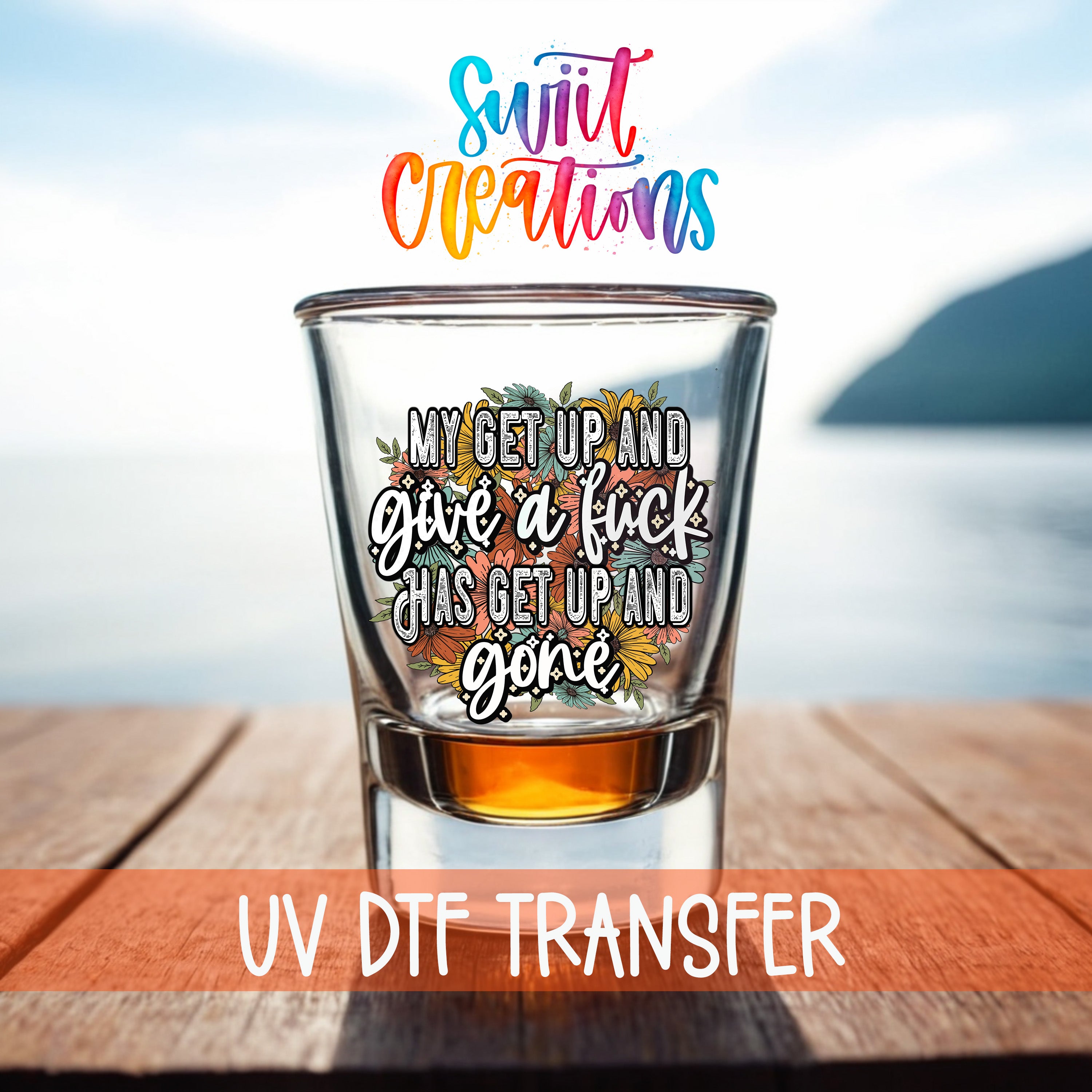 a shot glass with a colorful, artistic design and text on it, placed on a wooden surface with a body of water in the background.