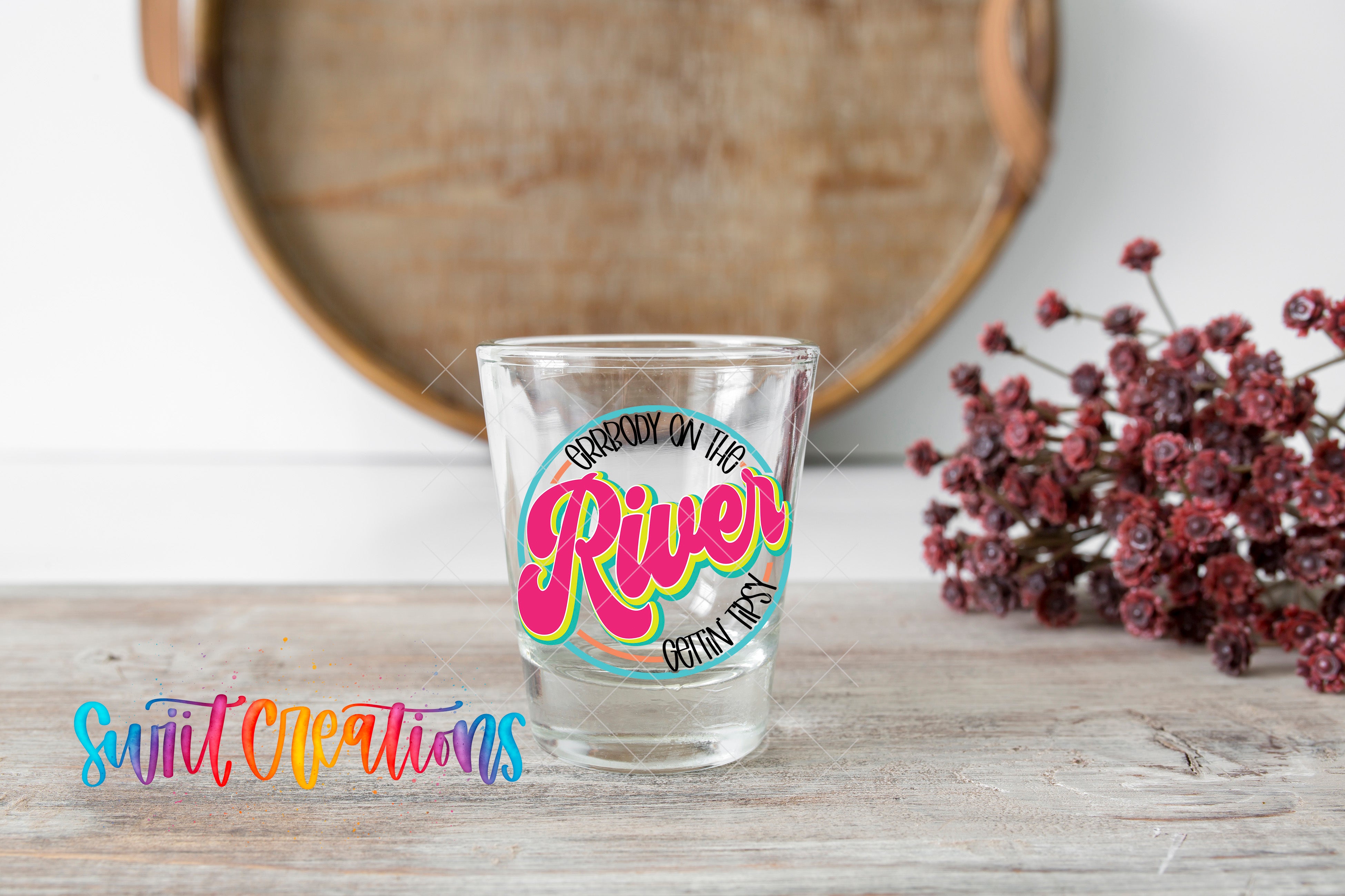 A shot glass with a colorful design sits on a wooden surface, with a wooden tray and a bouquet of red flowers in the background.