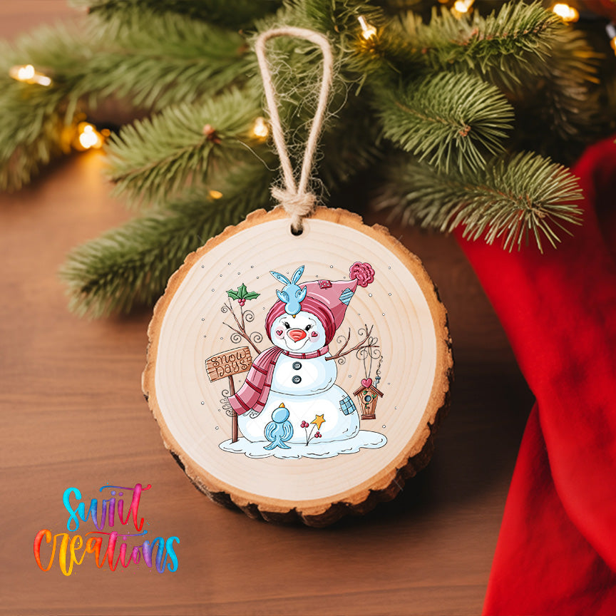 A wooden ornament featuring a snowman with a sign, surrounded by snow and twinkling lights, hangs on a Christmas tree.