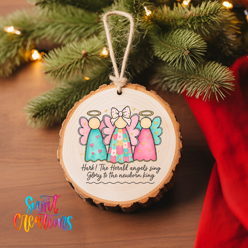 A wooden ornament with a design of three angels, each with a different color and style, hangs on a Christmas tree. The ornament is accompanied by a text that reads "Hark! The Herbed Angels sing glory to the newborn king".