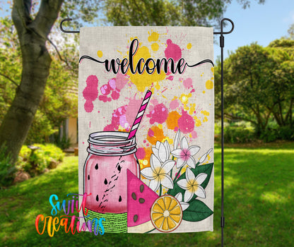 A colorful welcome sign with a mason jar, watermelon, and flowers, set against a backdrop of trees and greenery.