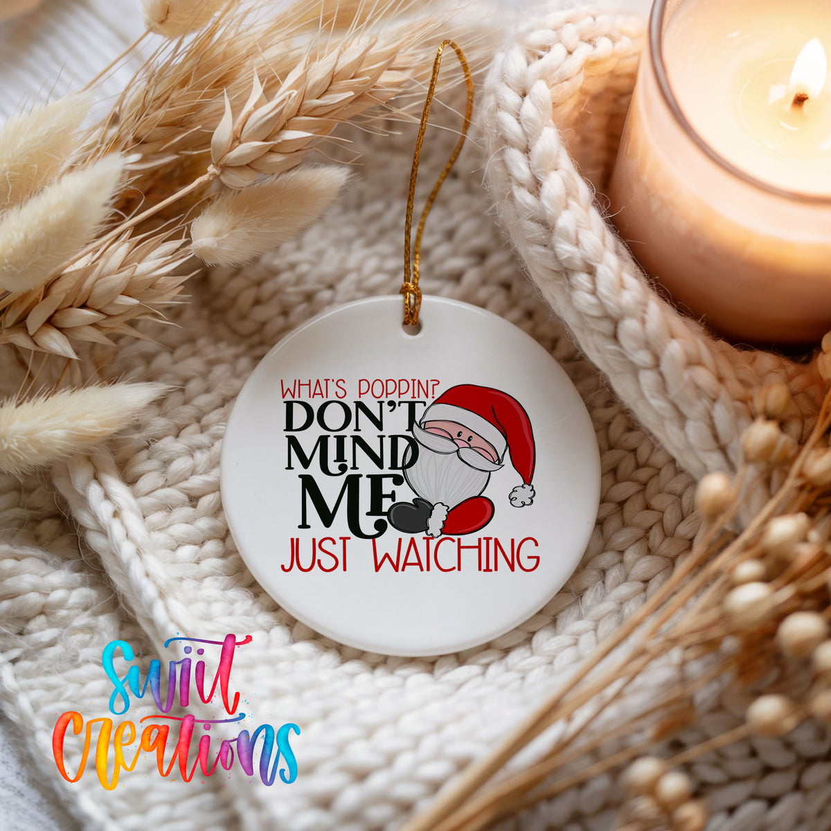 A white ornament with a Santa Claus design and text that says "What's Popping? Don't Mind Me Just Watching" is displayed on a knitted surface next to a lit candle.