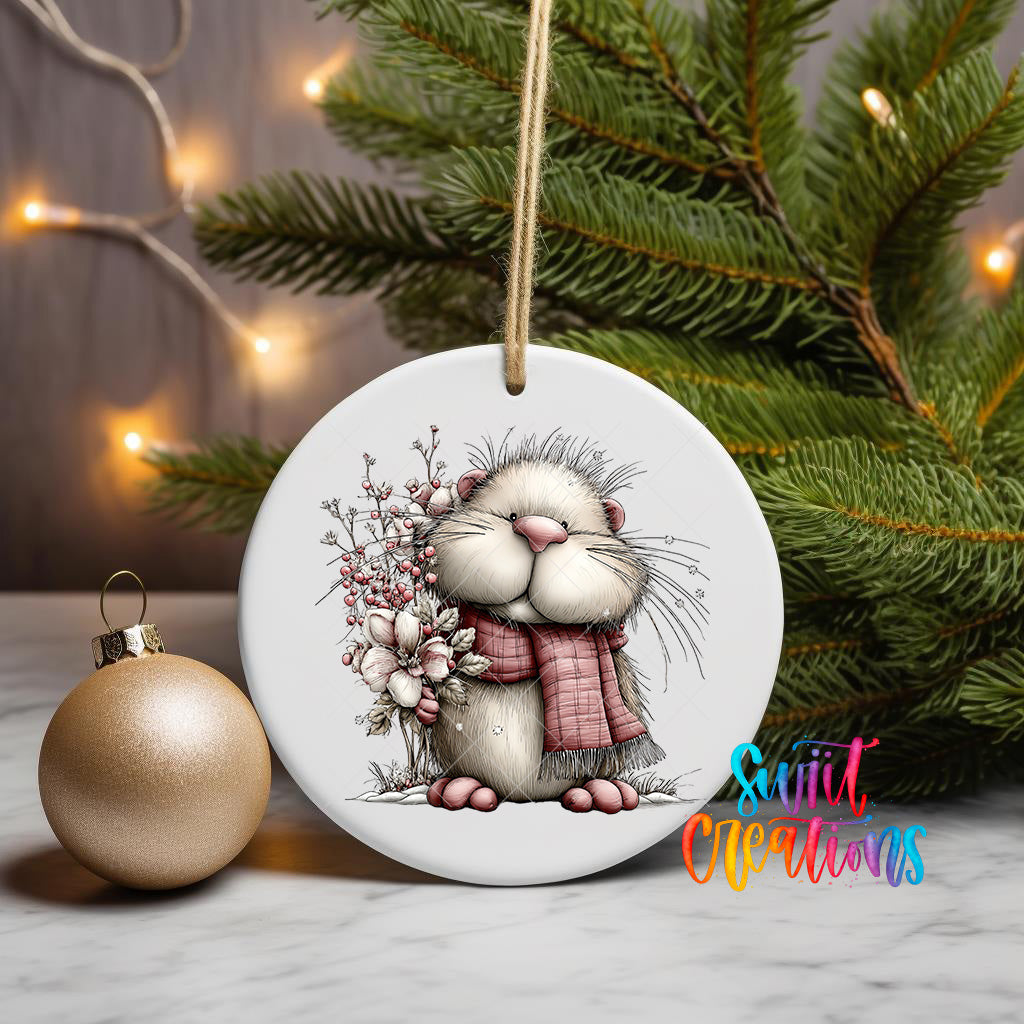 A white ceramic ornament featuring a cute cartoon rabbit wearing a red scarf and holding a flower, placed next to a gold ornament and a Christmas tree with twinkling lights.