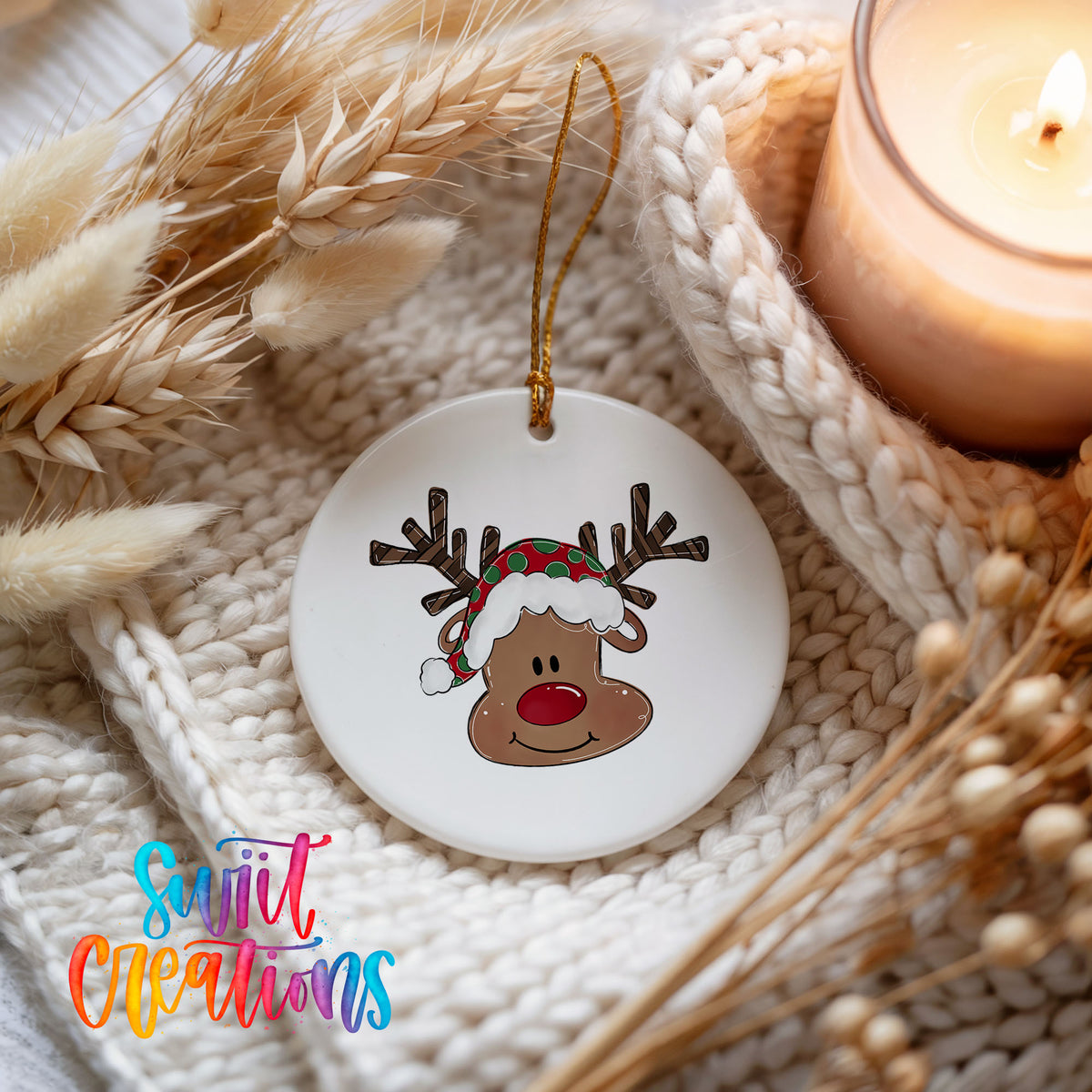A white ornament with a reindeer face and a Santa hat is displayed on a white knitted blanket, with a lit candle and dried flowers in the background.