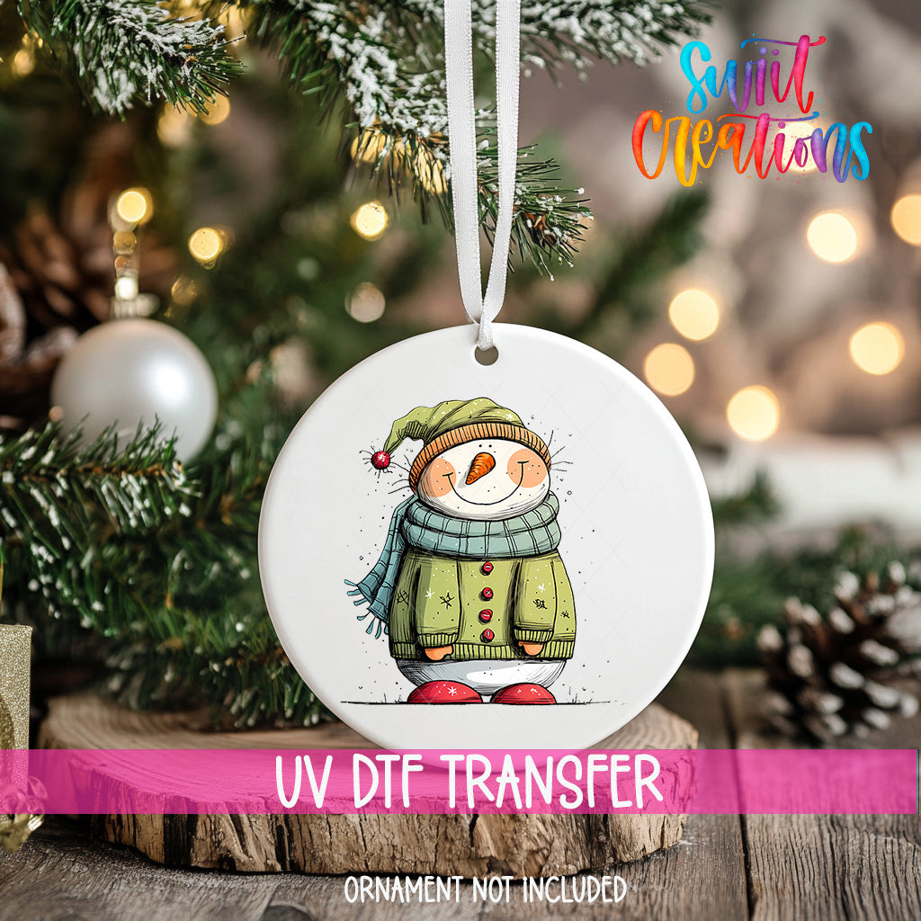 A white ornament with a snowman design hangs on a Christmas tree, surrounded by pine branches and twinkling lights.