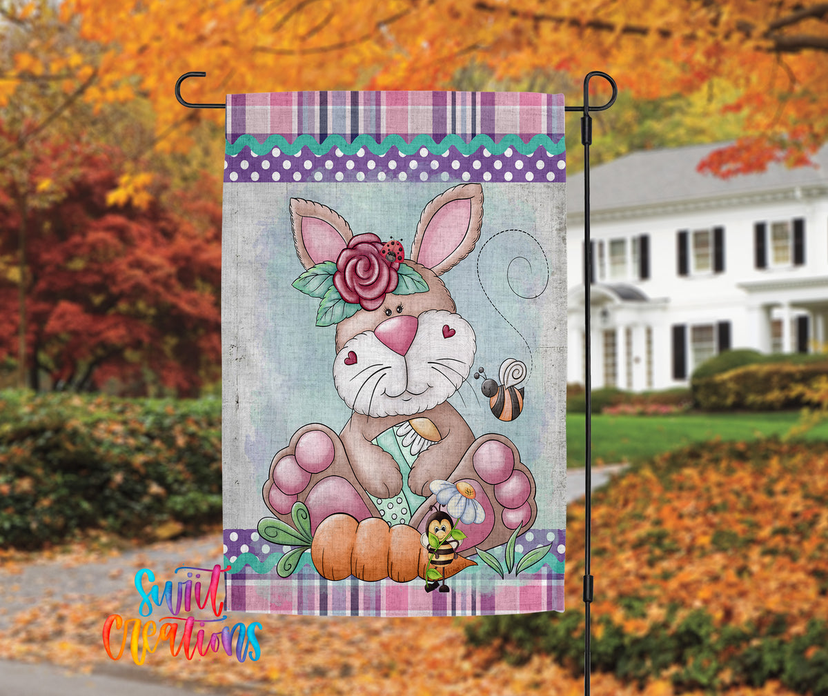 A colorful garden flag featuring a cartoon rabbit holding a carrot and a bee, with a house and autumn foliage in the background.
