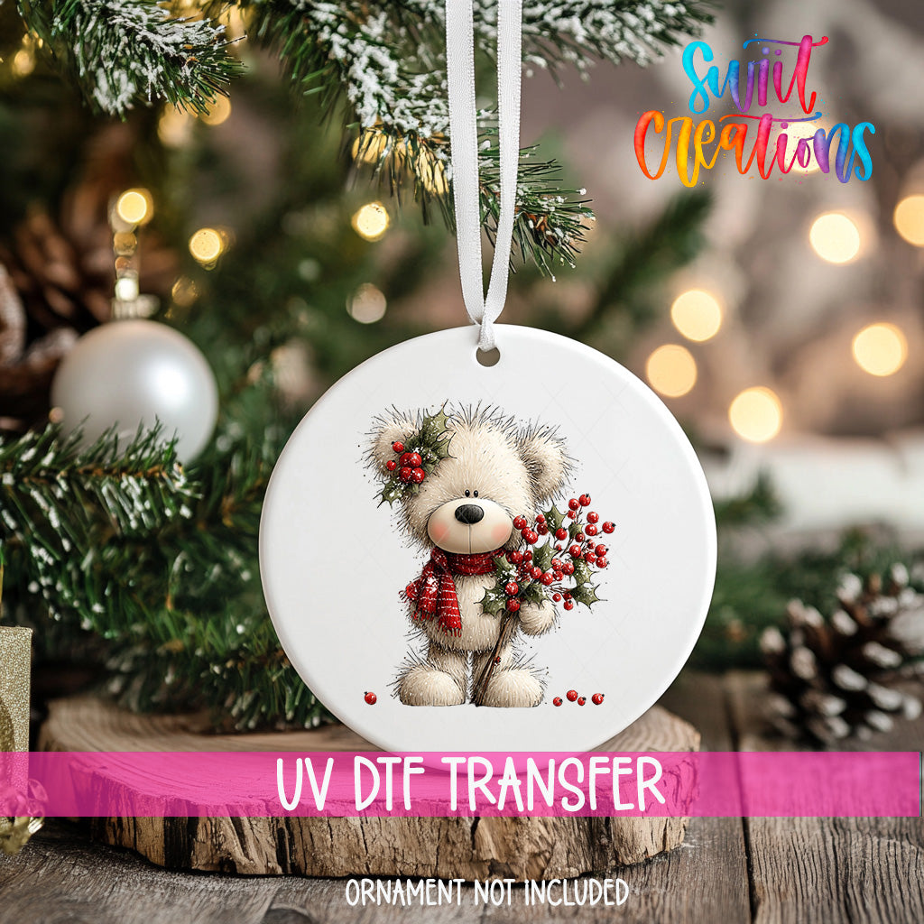 A white ornament with a teddy bear holding berries is hanging on a Christmas tree.