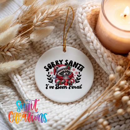 Sorry Santa I've Been Feral funny raccoon ceramic Christmas ornament styled beside a candle and cozy knit blanket
