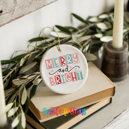 Round ceramic Christmas ornament featuring pastel block-letter Merry and Bright holiday typography, styled on books with holiday greenery