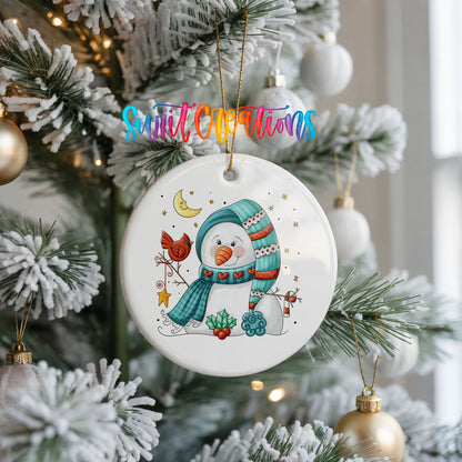 Round ceramic Christmas ornament featuring cozy snowman holding a bright red cardinal beneath a winter moon, hanging on a frosted Christmas tree