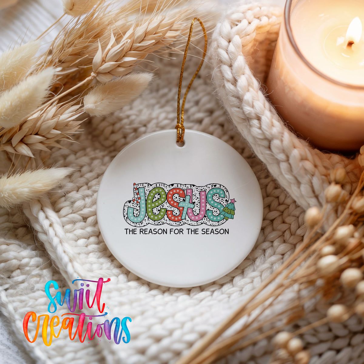 Round ceramic Christmas ornament featuring colorful Jesus Is the Reason for the Season typography, styled beside a candle and cozy knit blanket