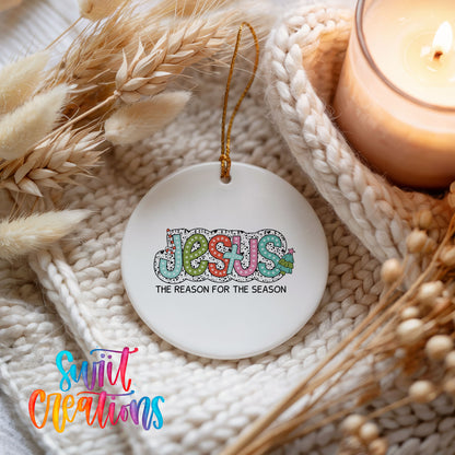 Round ceramic Christmas ornament featuring colorful Jesus Is the Reason for the Season typography, styled beside a candle and cozy knit blanket