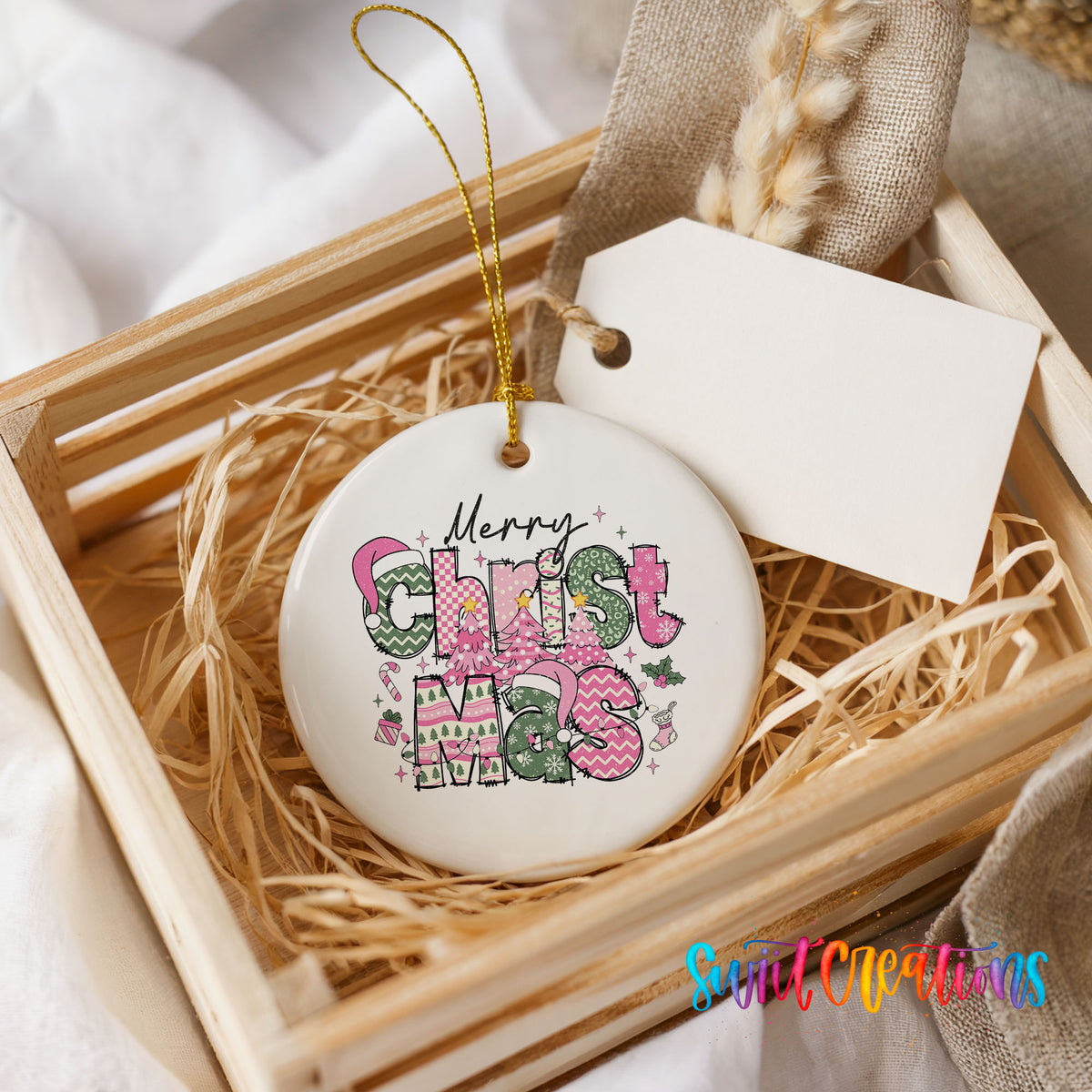 Round ceramic Christmas ornament featuring stacked patterned Merry Christmas lettering in pink green and black, presented in a rustic wooden gift crate