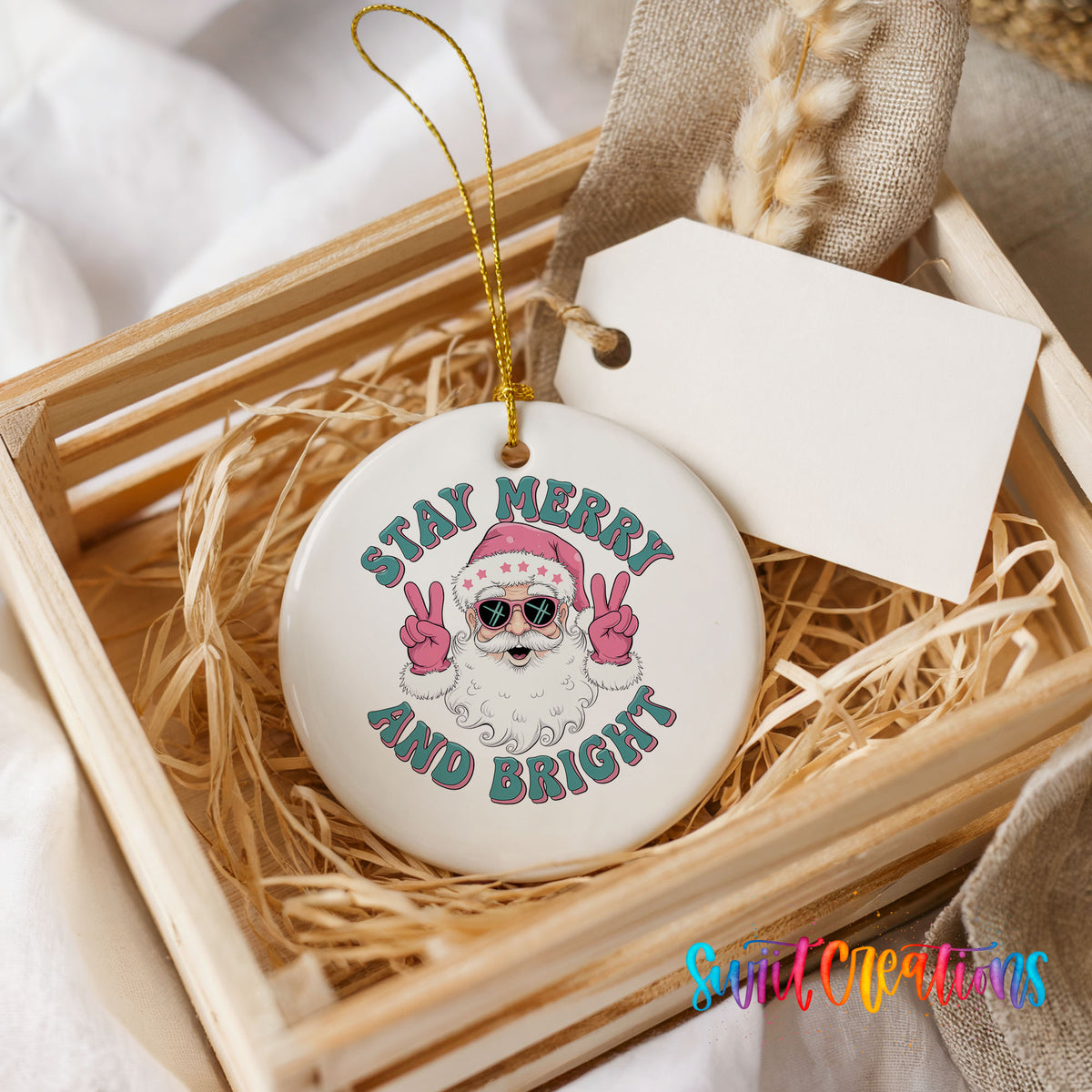 Round ceramic Christmas ornament featuring Santa in sunglasses giving peace signs with Stay Merry and Bright lettering, presented in a rustic wooden gift crate