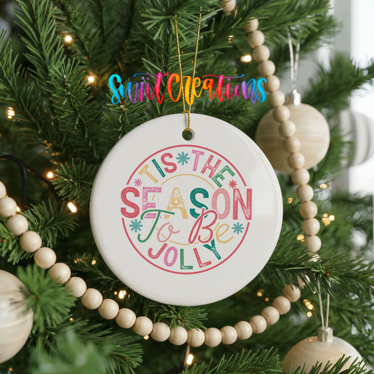 Round ceramic Christmas ornament featuring round pastel ’Tis the Season to Be Jolly holiday typography, hanging on a decorated Christmas tree