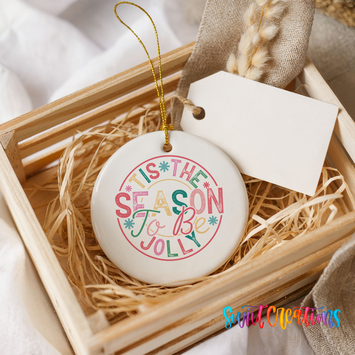 Round ceramic Christmas ornament featuring round pastel ’Tis the Season to Be Jolly holiday typography, presented in a rustic wooden gift crate