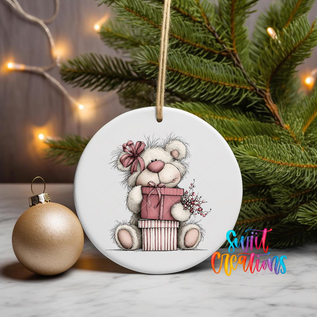 A white ceramic ornament featuring a teddy bear holding a gift box, surrounded by a Christmas tree and twinkling lights.