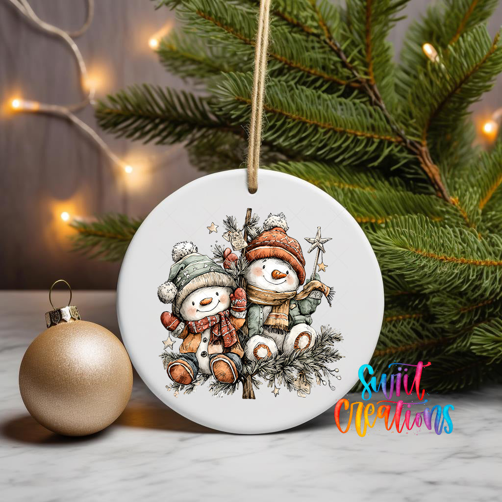 A white ceramic ornament featuring two snowmen is displayed on a table, with a gold ornament and a Christmas tree in the background.