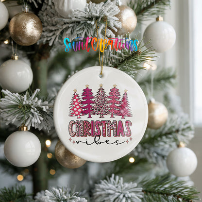 A white Christmas ornament with a pink and leopard print design hangs from a tree, surrounded by white and gold ornaments.
