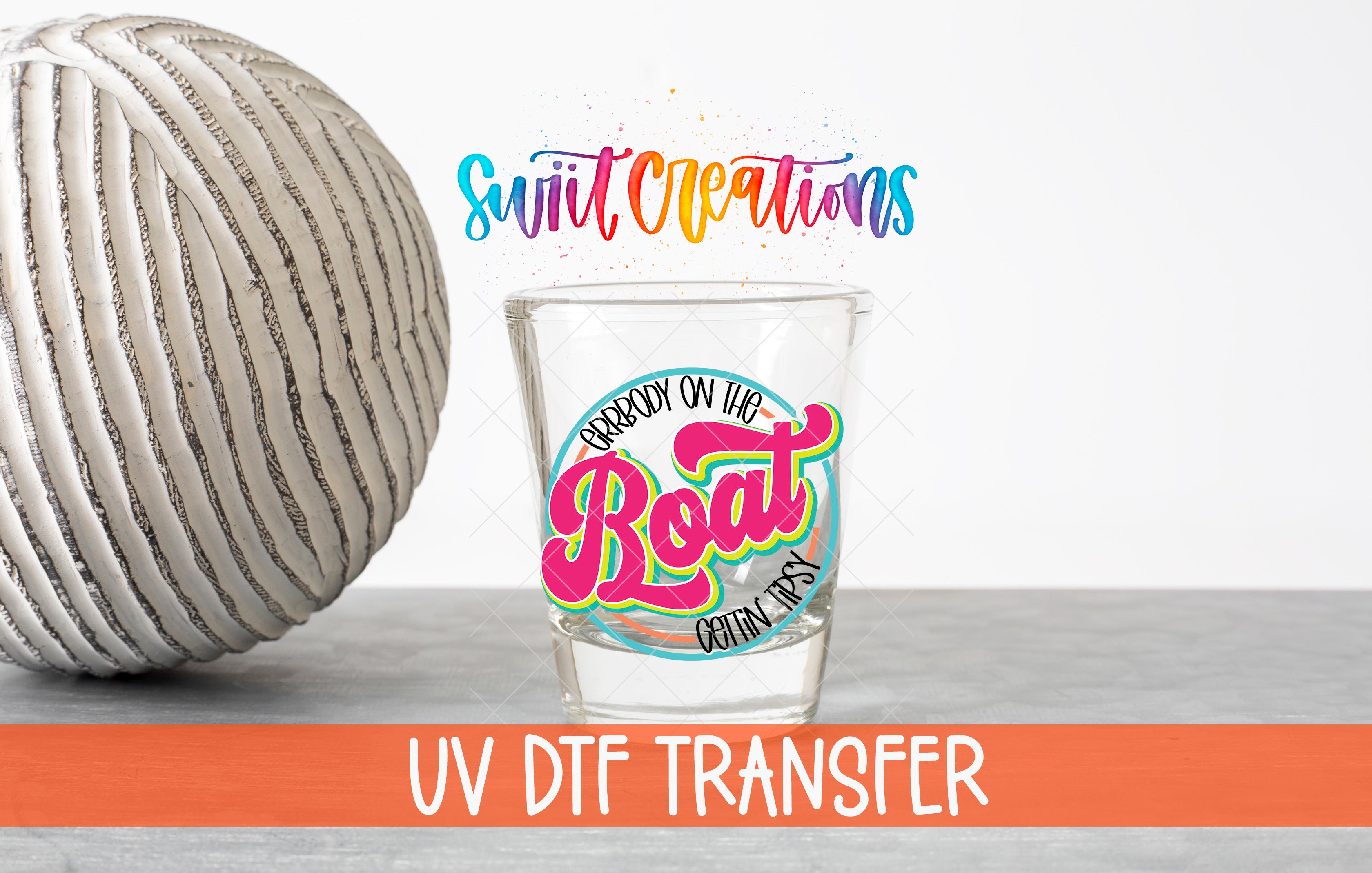 a glass with a colorful design and text, along with a gray ball and a striped object, all placed on a gray surface.