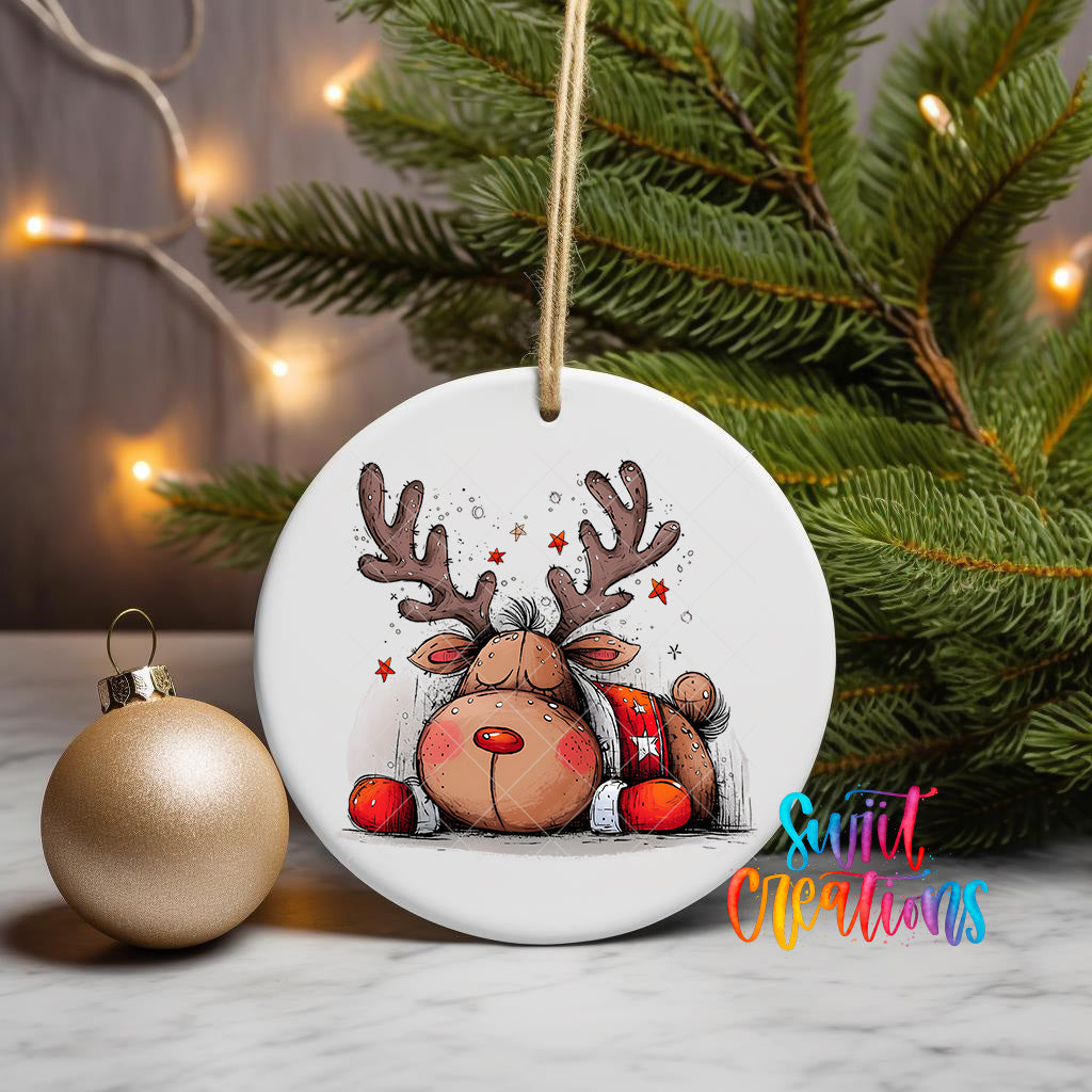 A white ceramic ornament featuring a cartoon reindeer with antlers and a red bow, hanging on a string next to a gold ornament and a Christmas tree.