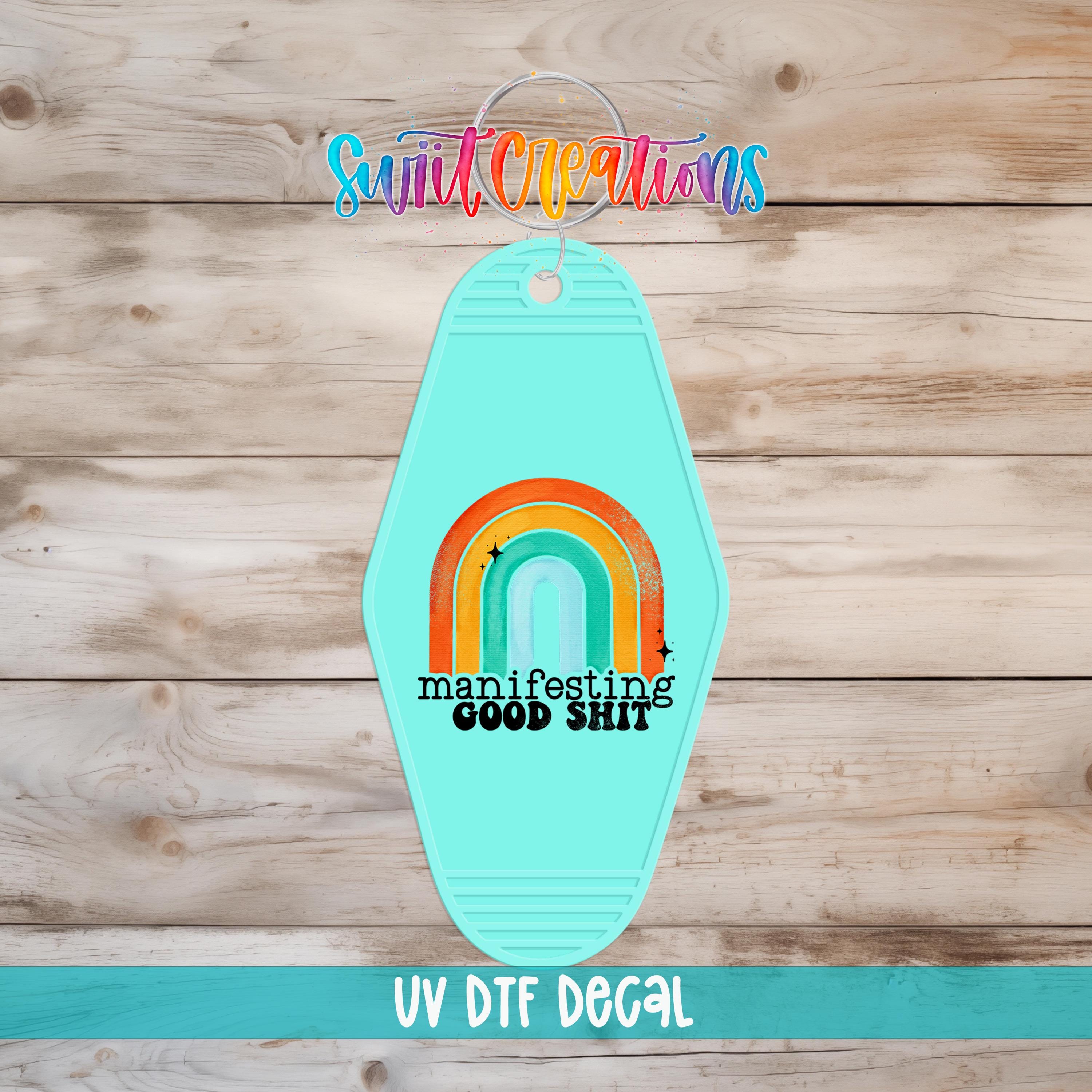 A turquoise-colored keychain with a rainbow graphic and the text "manifesting GOOD SHIT" printed on it, along with the text "UV Dtf Decal" at the bottom.