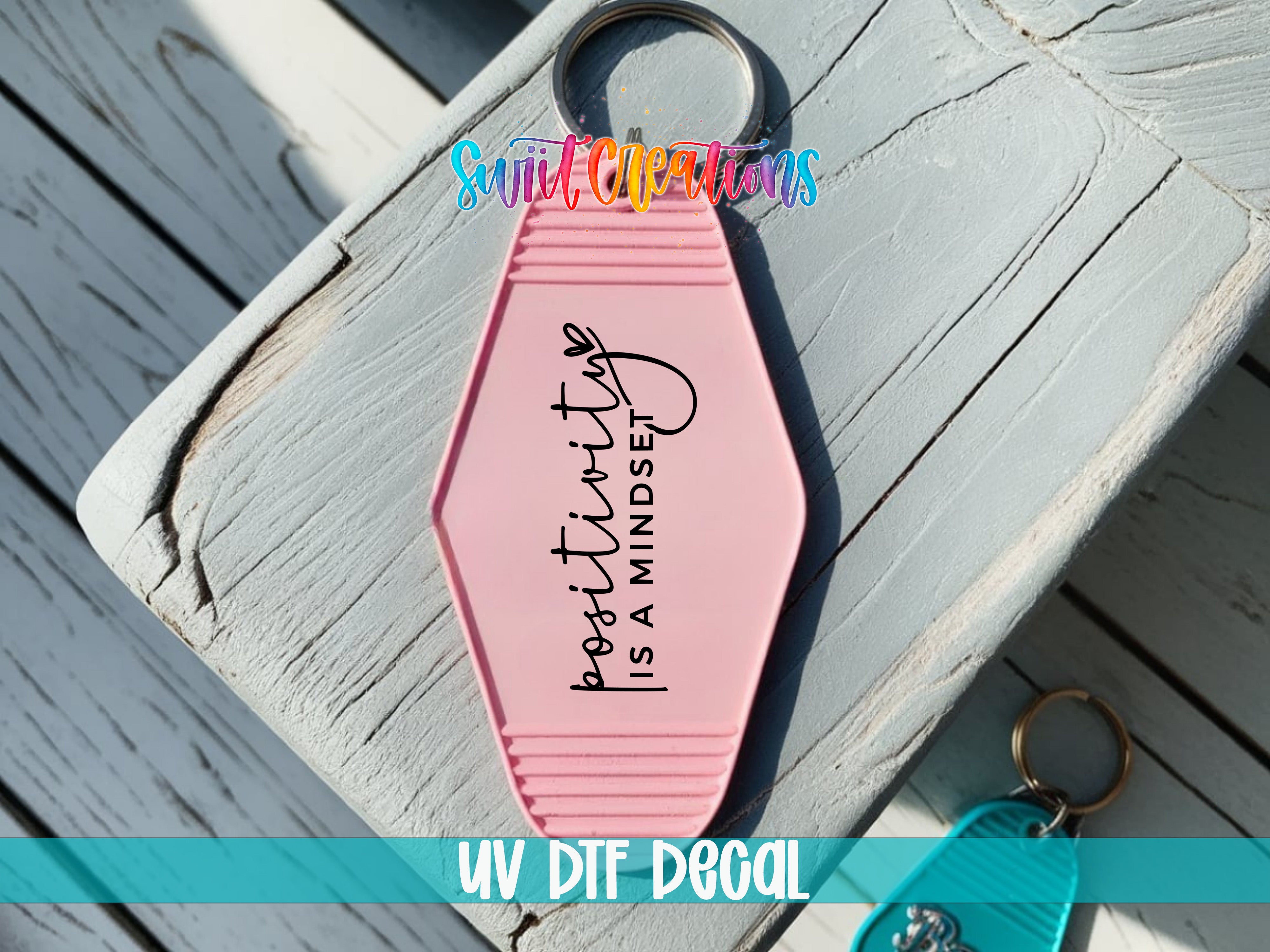 A pink keychain with the text "positivity is a mindset" printed on it, placed on a wooden surface.