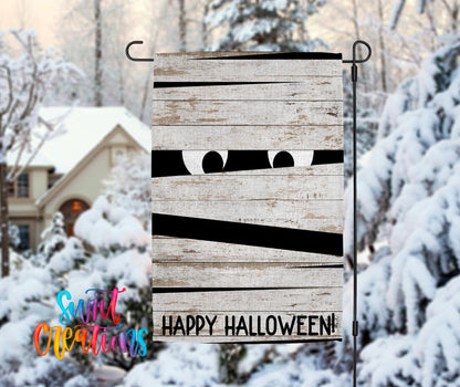 A Halloween-themed wooden sign with a mummified face peeking out from behind a wooden plank, set against a snowy winter landscape with a house in the background.