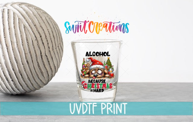 a shot glass with a Christmas-themed design and text, placed next to a white ball on a gray surface.