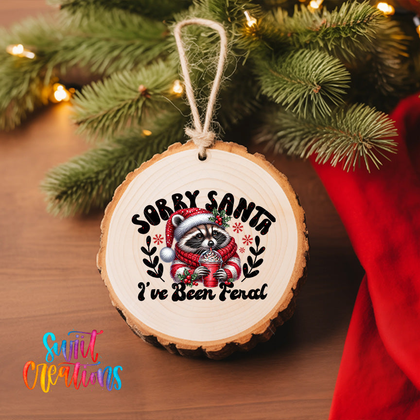 A wooden ornament with a raccoon wearing a Santa hat and scarf, and the text "Sorry Santa, I've been fired" is hanging on a Christmas tree.