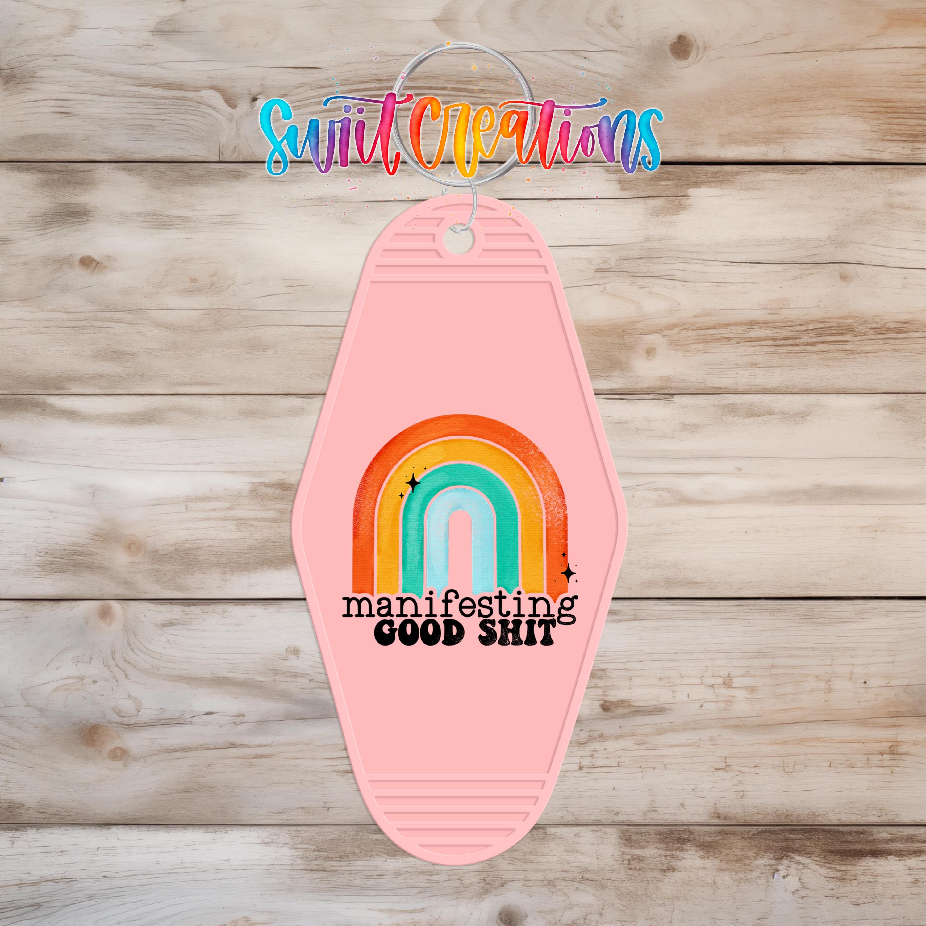 A pink keychain with a rainbow graphic and the text "manifesting good shit" printed on it, hanging on a wooden surface.