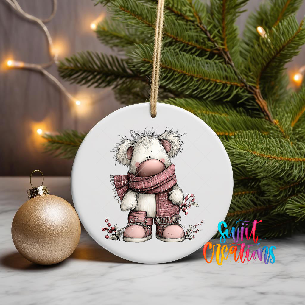 A white ceramic ornament featuring a cartoon image of a bear wearing a red scarf and boots, hanging on a string next to a gold ornament and a Christmas tree.