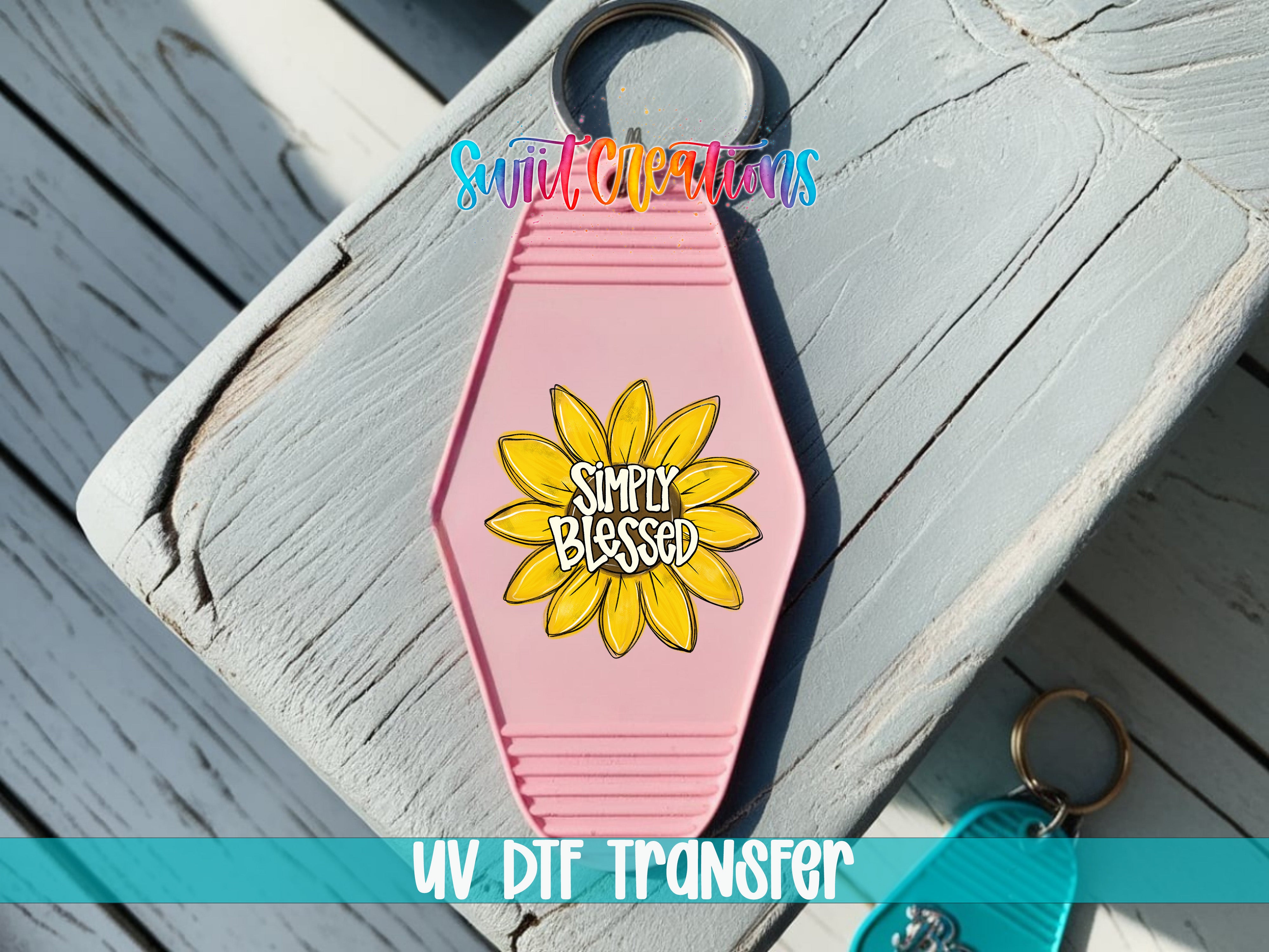 A pink keychain with a sunflower design and the text "Simply Blessed" printed on it, placed on a wooden surface.