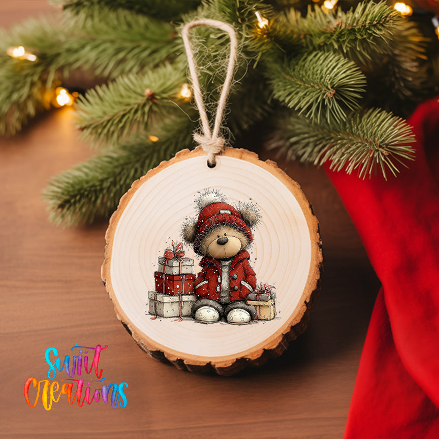 A wooden ornament featuring a teddy bear wearing a red coat and hat, sitting next to a stack of presents, is hanging on a Christmas tree.
