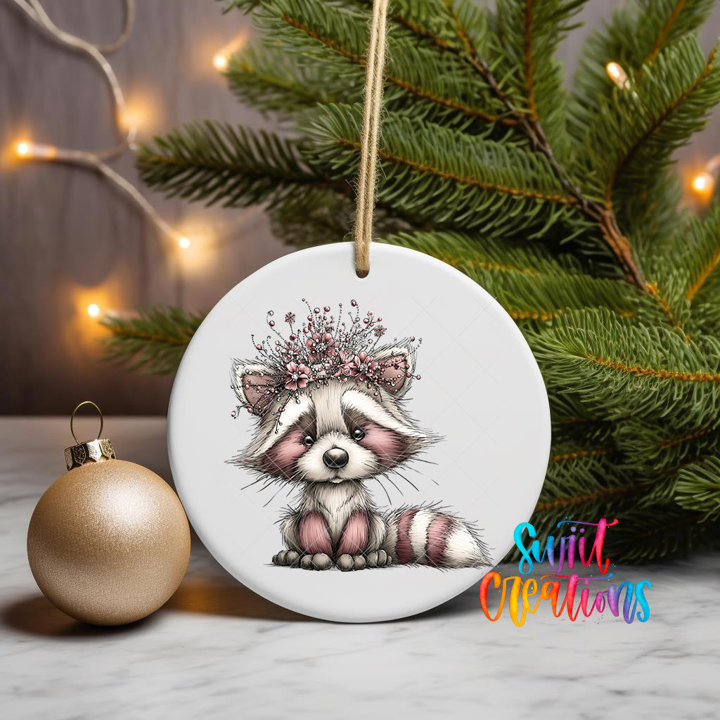 A white ceramic ornament featuring a cute raccoon wearing a floral crown, placed on a marble surface next to a gold ornament and a Christmas tree with twinkling lights.