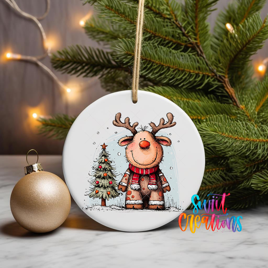 A white ceramic ornament featuring a cartoon reindeer wearing a red scarf and standing next to a Christmas tree, with a gold ornament and a string of lights in the background.