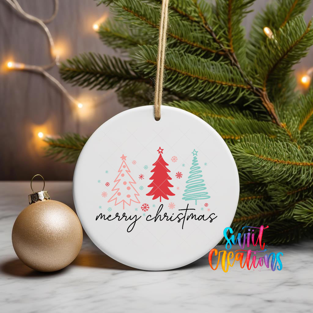 A white ceramic ornament with the text "Merry Christmas" and three stylized Christmas trees is displayed on a marble surface, with a gold ornament and a pine tree in the background.