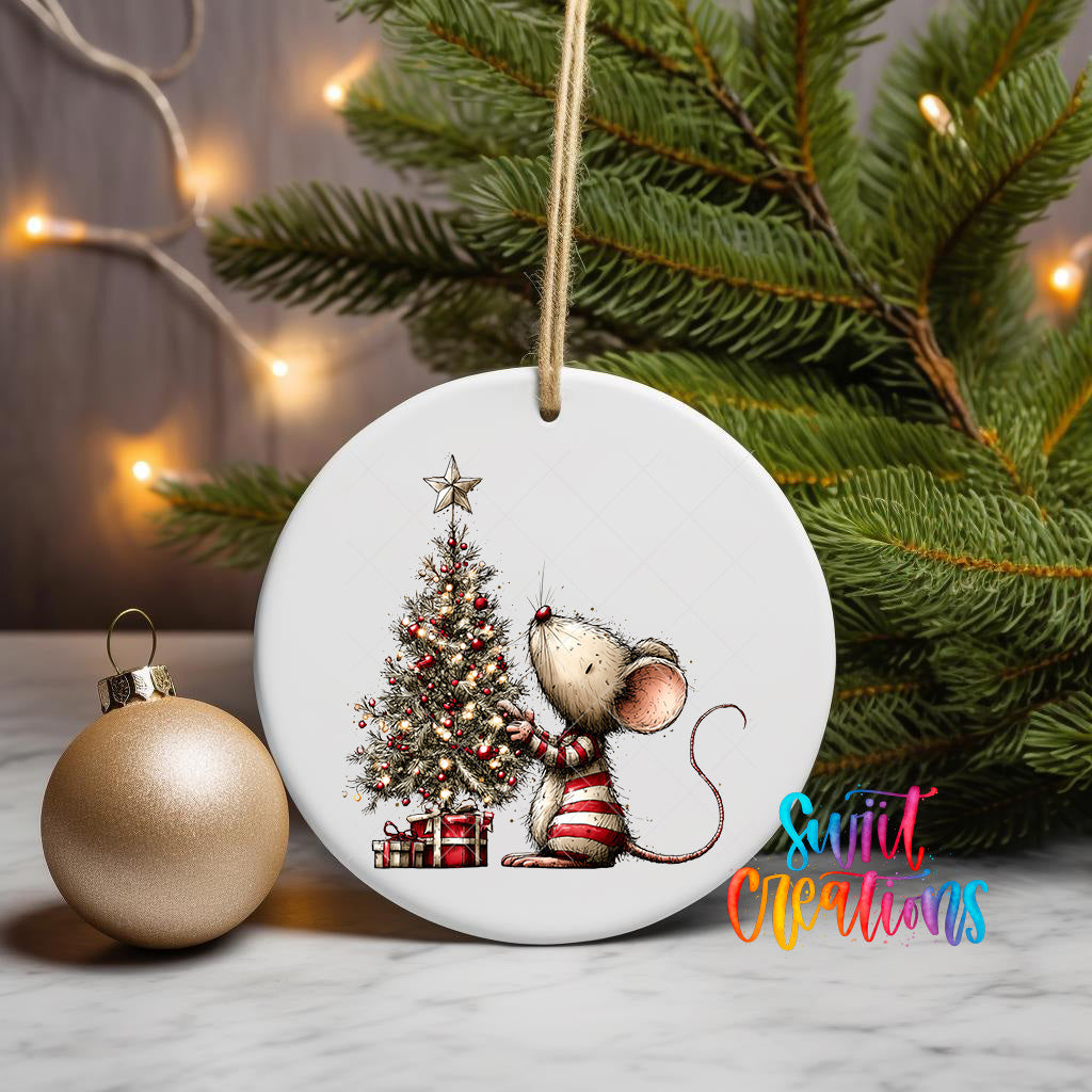 A white ceramic ornament featuring a cartoon mouse sitting next to a Christmas tree, with a star on top of the tree. The ornament is hanging from a string, and there is a gold ornament and a Christmas tree in the background.