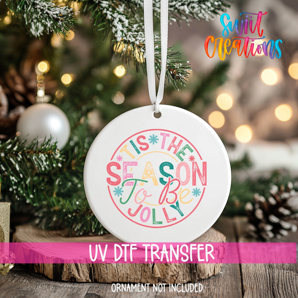 A white ceramic ornament with a colorful design hangs on a Christmas tree, surrounded by pine branches and other holiday decorations.