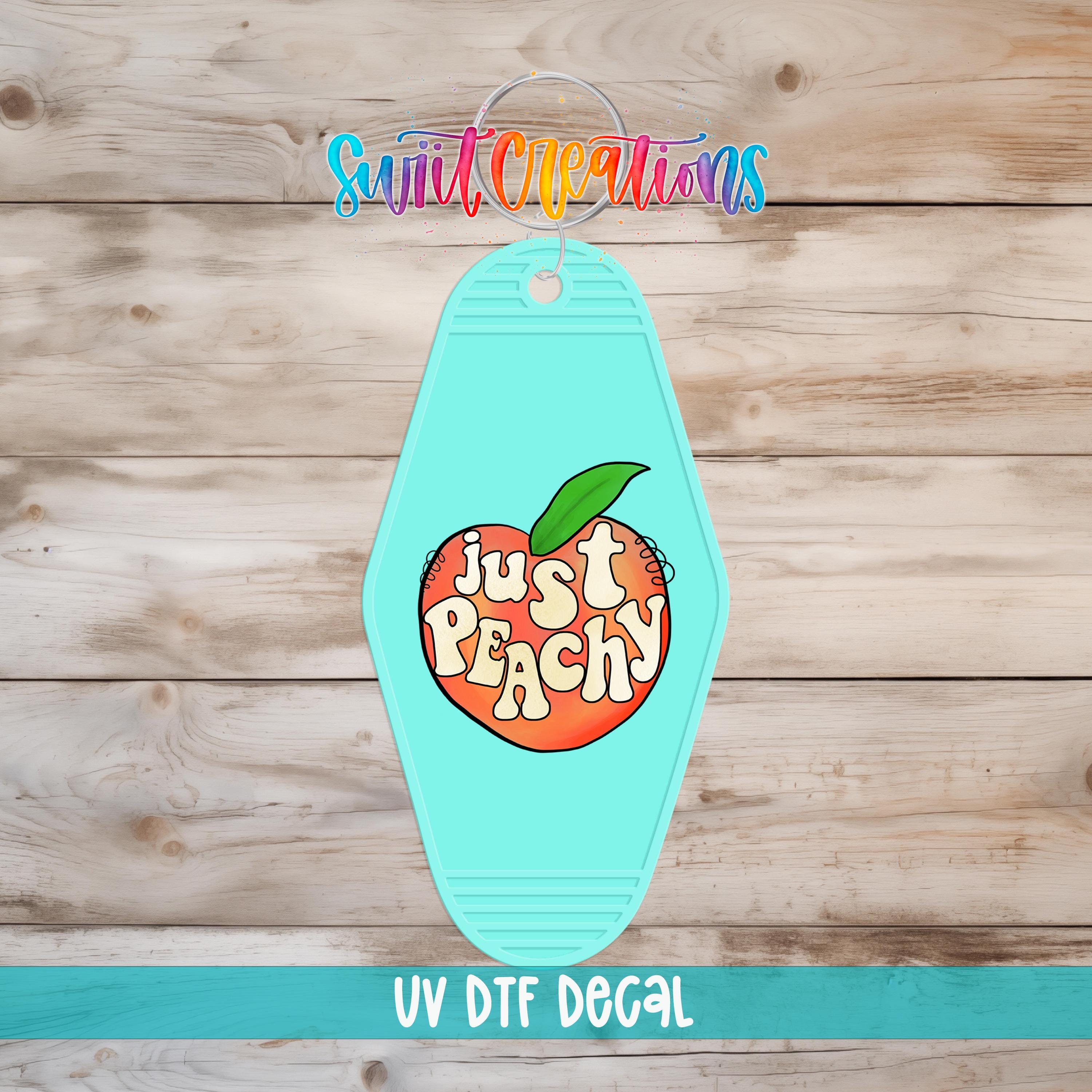 A turquoise keychain with the text "Just Peachy" and an illustration of a peach on it, placed on a wooden surface.