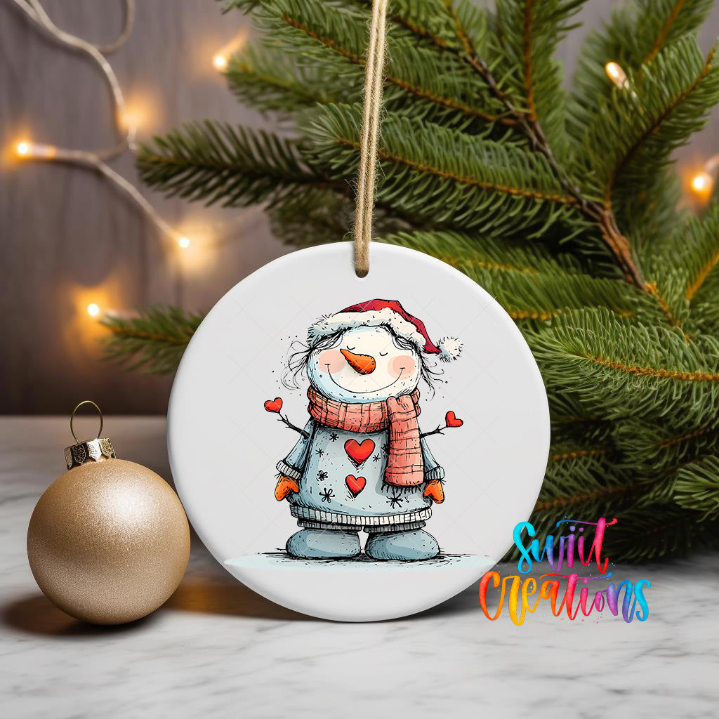 A white ceramic ornament featuring a smiling snowman wearing a red scarf and hat, with hearts on its arms, is displayed on a white surface next to a gold ornament and a Christmas tree.