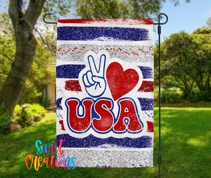 A patriotic flag with the word "USA" prominently displayed, featuring a peace sign and a red heart symbol. The flag is hanging outdoors in a grassy area with trees in the background.