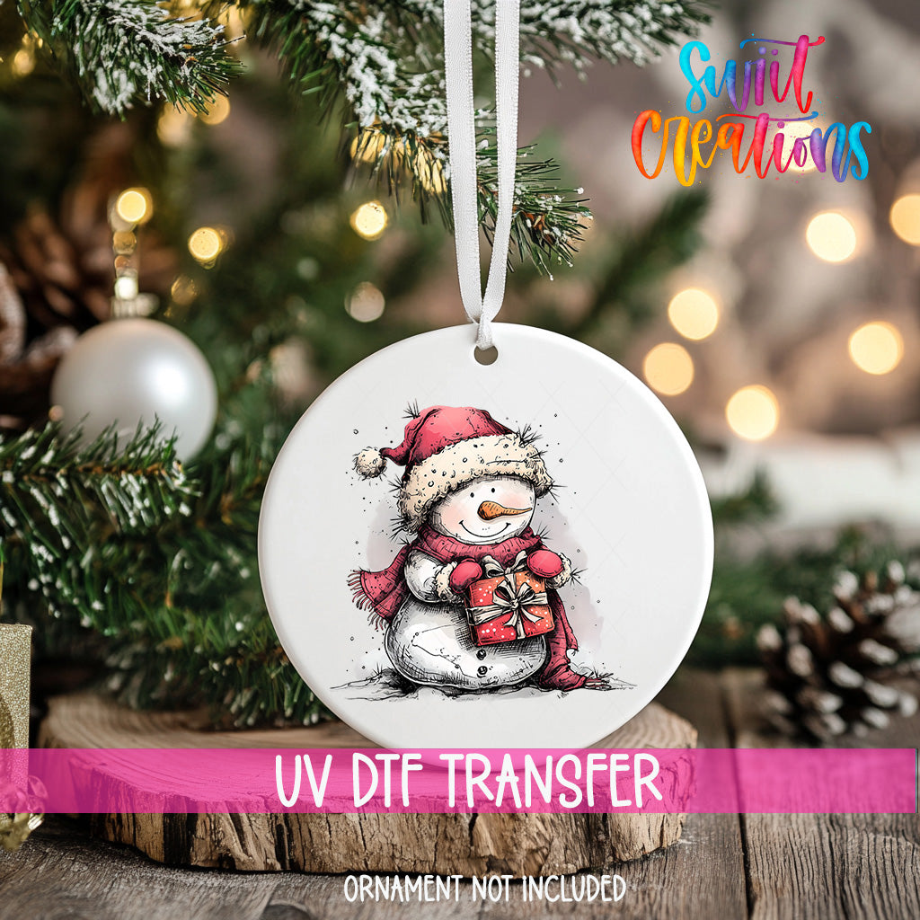 A white ornament with a snowman design is hanging on a Christmas tree, with a blurred background of pine branches and twinkling lights.