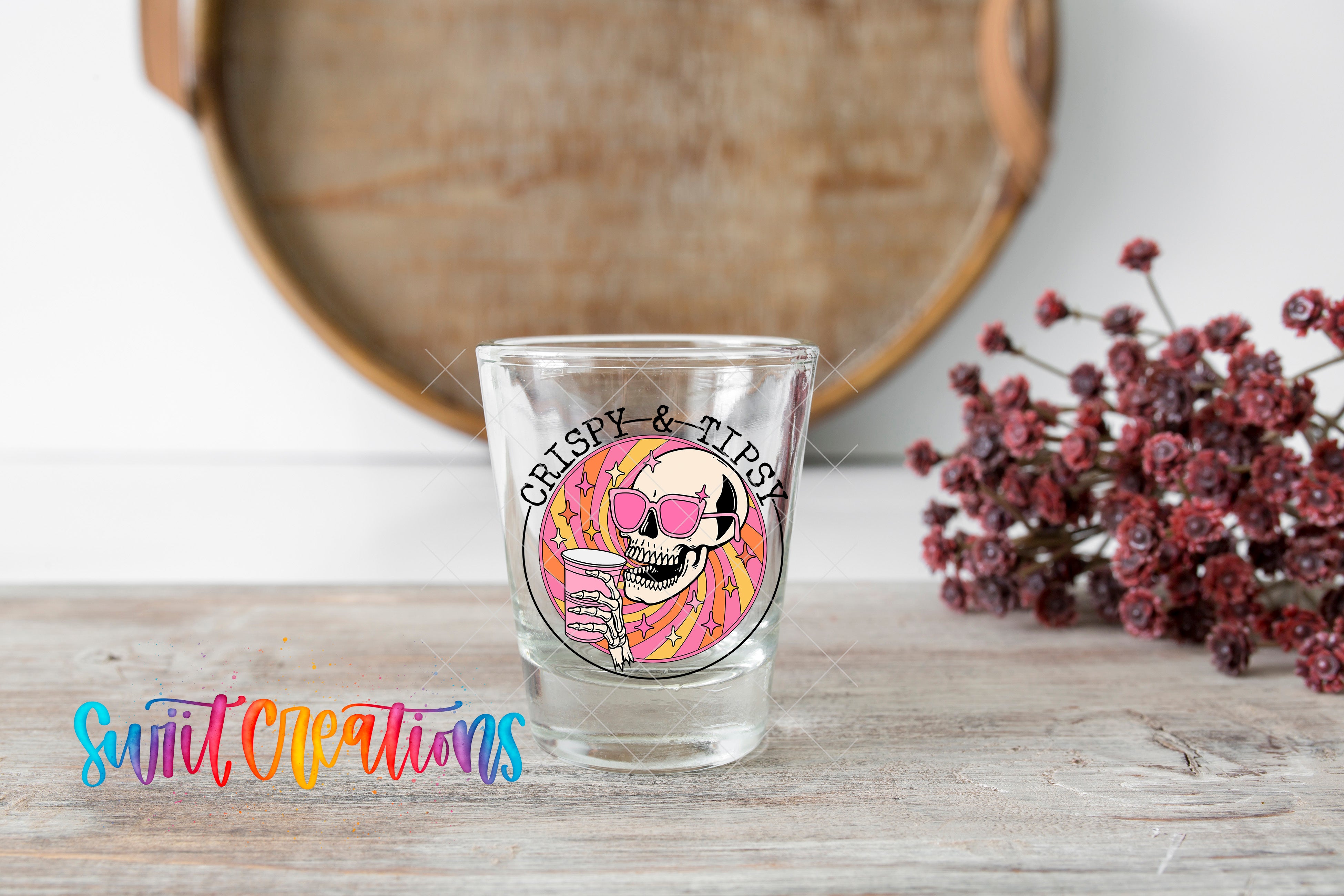 A shot glass with a skull and sunglasses design, placed on a wooden surface next to a bunch of red flowers.