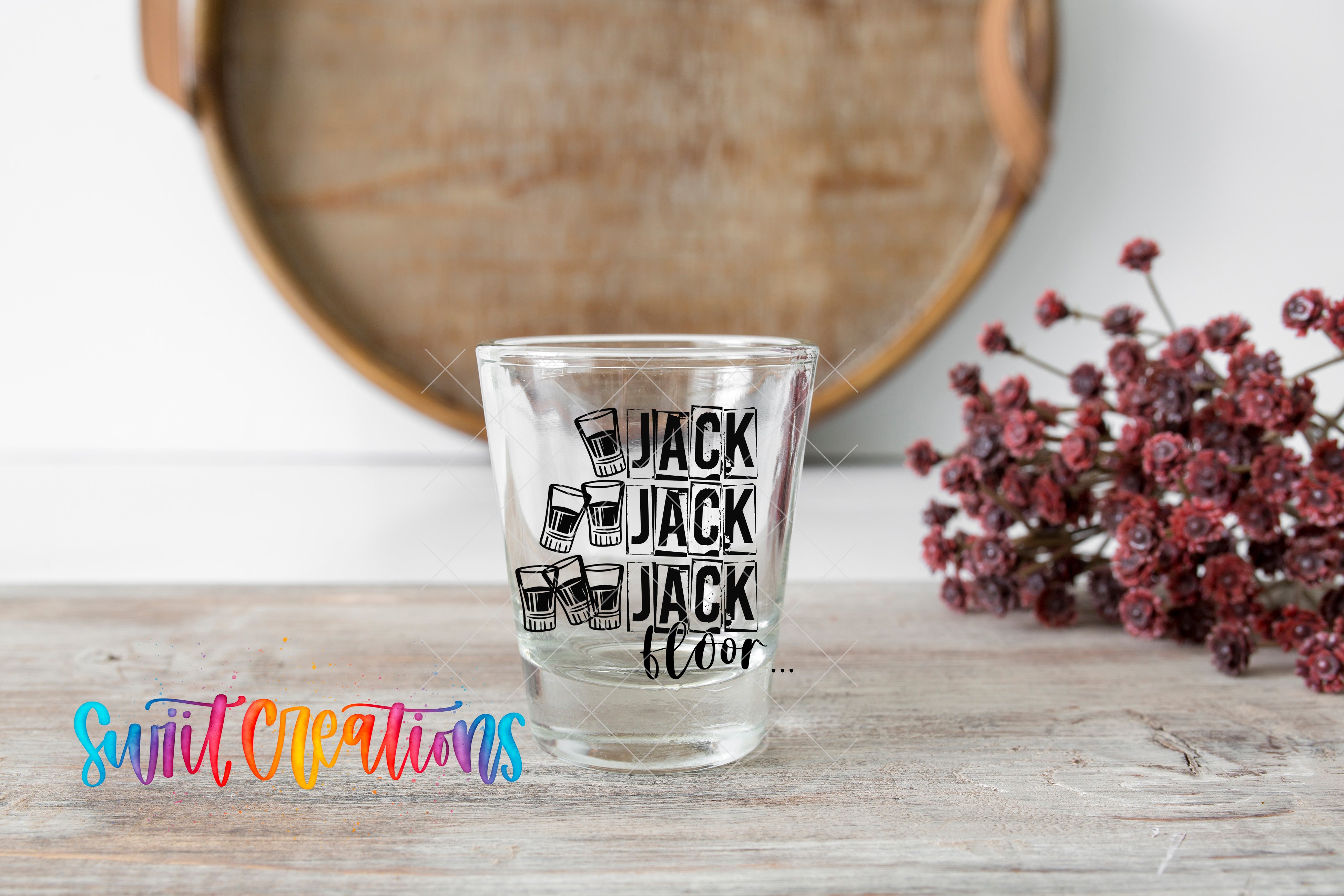 A shot glass with the text "JACK JACK JACK JACK JACK" printed on it, placed on a wooden surface next to a bunch of red flowers.