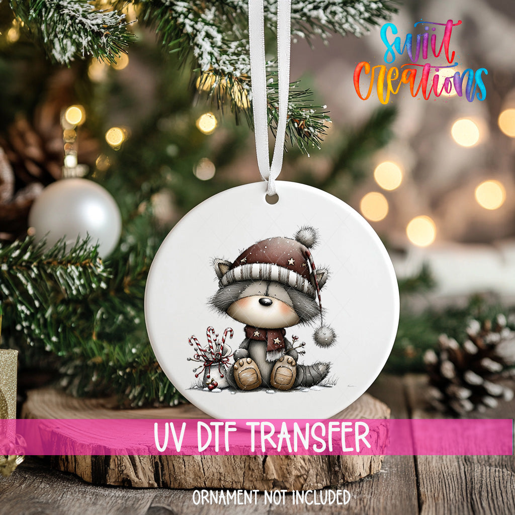 A white ceramic ornament with a cartoon raccoon wearing a hat and holding a small bag is hanging on a Christmas tree.