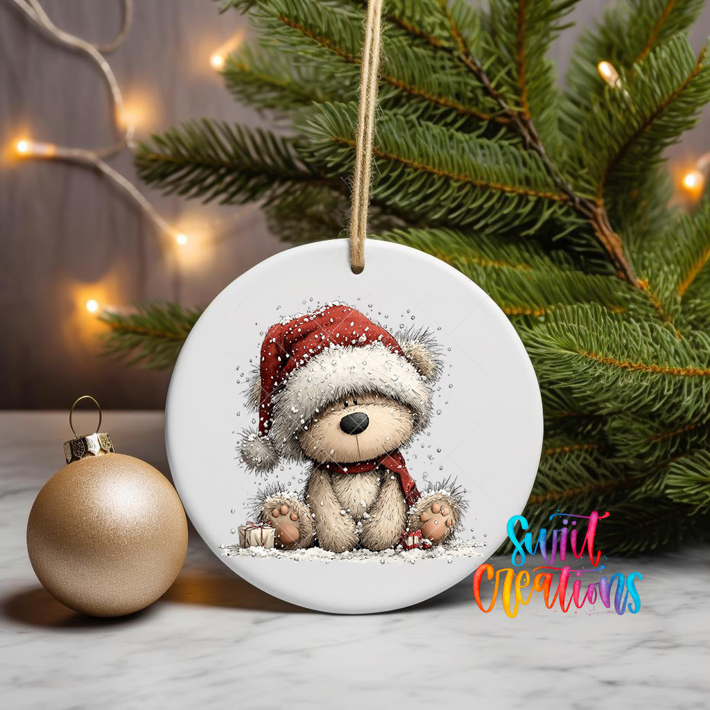 A white ceramic ornament featuring a teddy bear wearing a Santa hat and scarf, sitting on a snowy surface, with a Christmas tree and twinkling lights in the background.