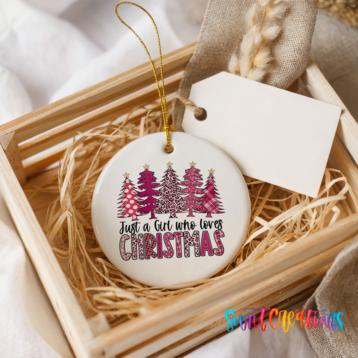 A white ceramic ornament with a Christmas tree design and the text "Just a Girl who loves Christmas" is displayed in a wooden crate.