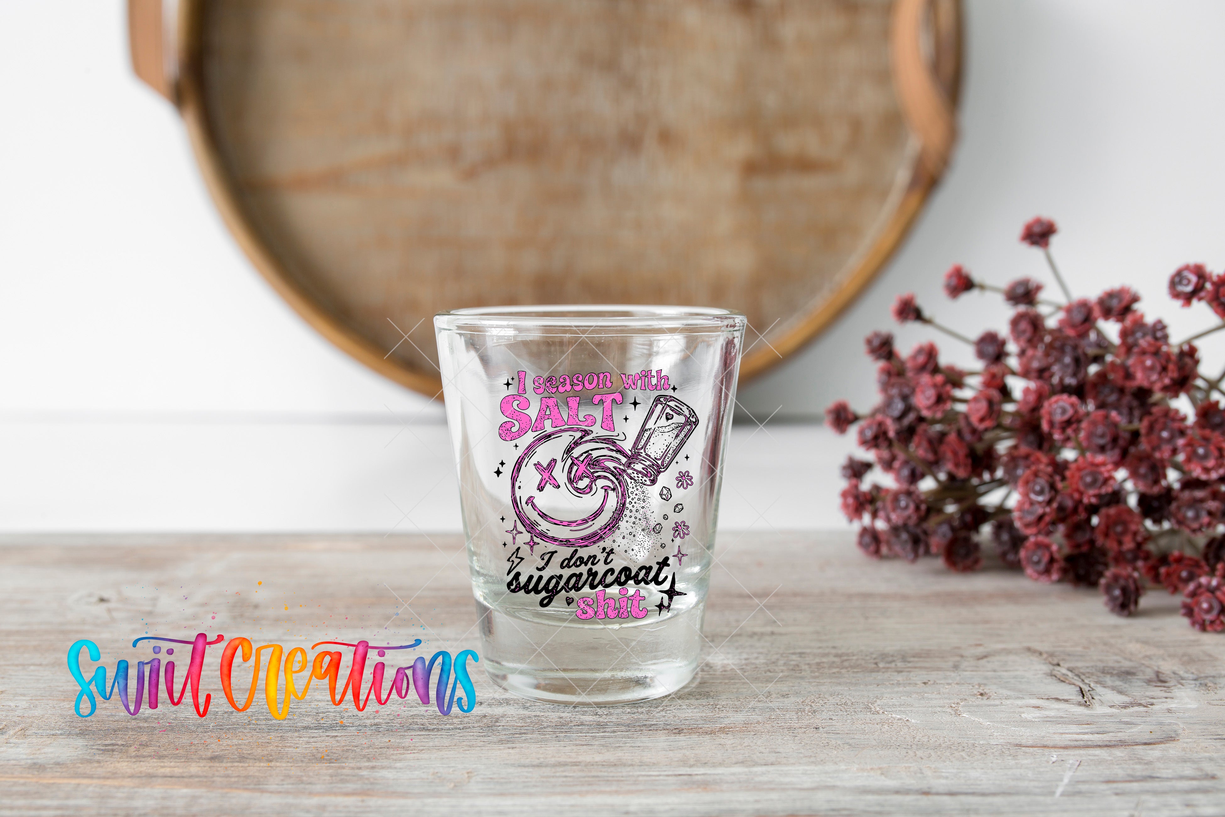 A shot glass with a colorful design and text on it, placed on a wooden surface next to a bunch of red flowers.