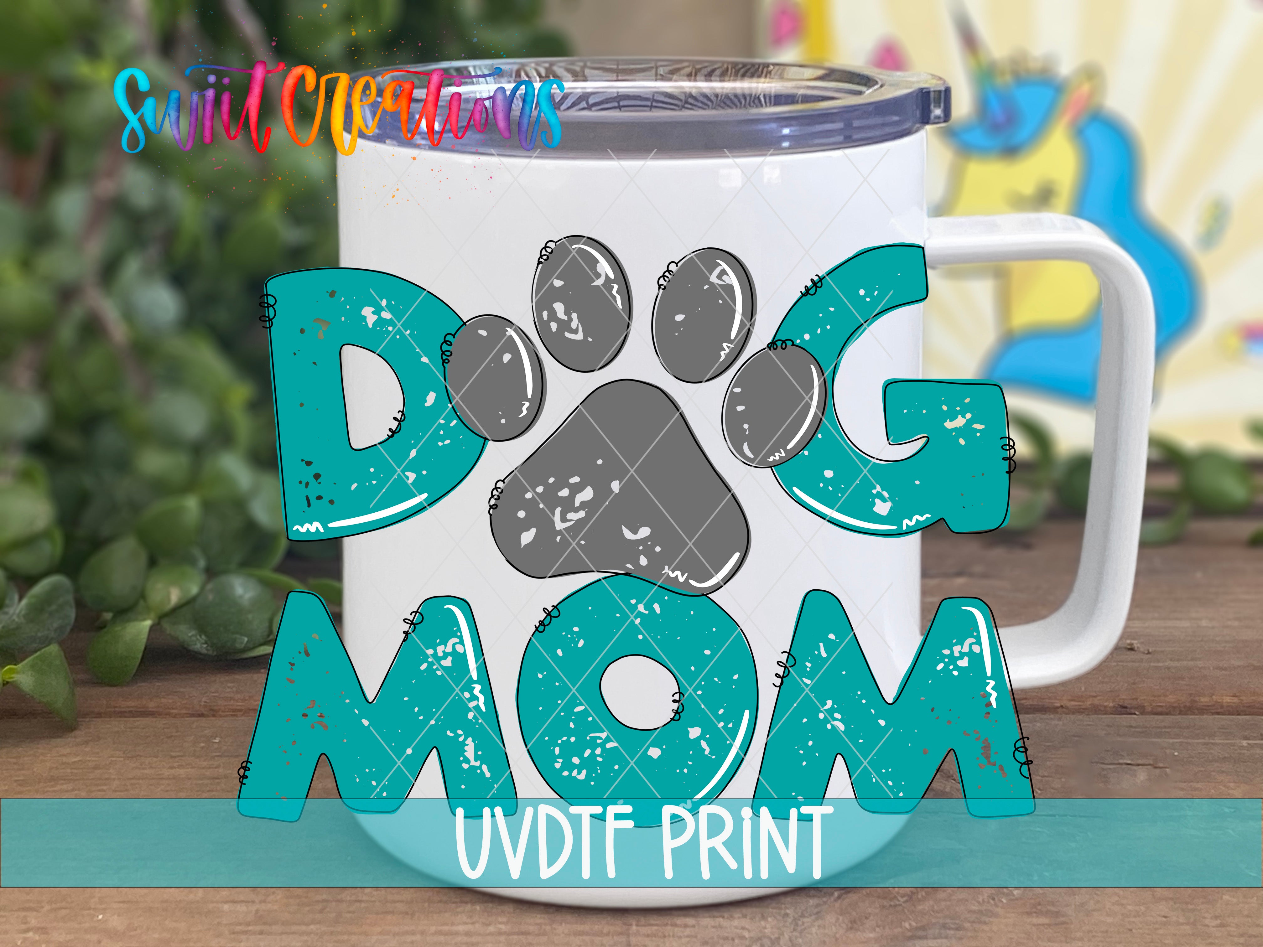A white mug with the text "DOG MOM" printed on it, along with a paw print design. The mug is placed on a wooden surface, and there is a blurred background that includes some greenery.